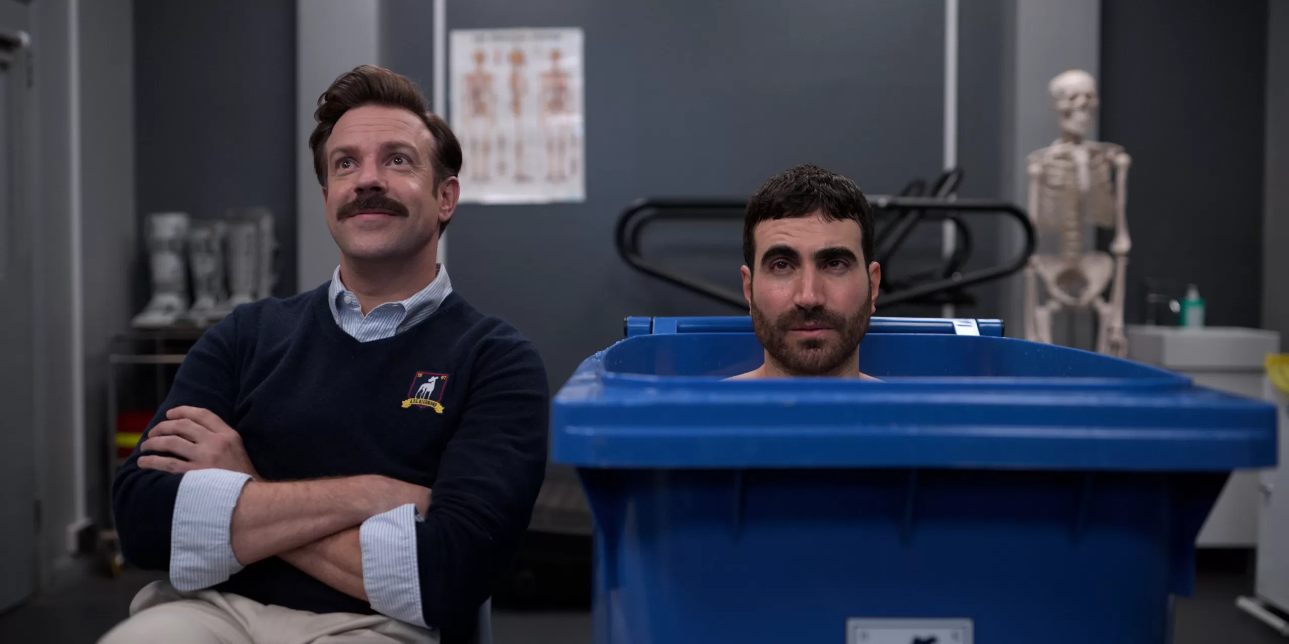 Ted Lasso and Roy Kent in the changing room. Roy is sat in a wheely bin.
